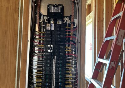 An open electrical panel with numerous wires and circuit breakers is mounted on wooden framing. A red ladder stands nearby.
