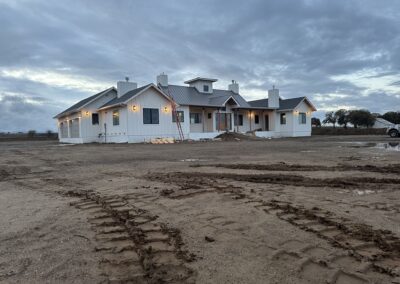 A newly constructed, single-story white house with a metal roof sits on a dirt lot under a cloudy sky.