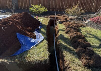 A dug trench with exposed soil and a partially buried pipe in a backyard, surrounded by grass and a wooden fence.