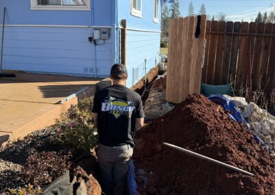A worker in a black shirt is standing in a trench next to a blue house. Dirt piles are on either side, and a wooden fence is nearby.