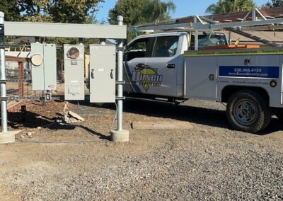 Utility truck parked beside electrical equipment on a gravel area, surrounded by trees and a clear sky.