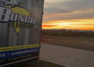 A trailer with "Bunch Electric" on the side is parked near a road. In the background, there's a sunset over a grassy field.