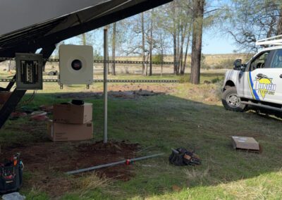 Solar panel installation in a grassy area near trees, with a utility vehicle parked nearby. A box and tool bag are on the ground.