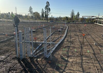 Construction site with pipes and rebar grids laid out on a level dirt surface under a clear sky. Sparse trees and utility poles are visible in the background.